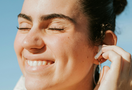 A smiling person with their eyes closed is wearing a white wireless earbud in their left ear. They are outside on a sunny day with a clear blue sky in the background.