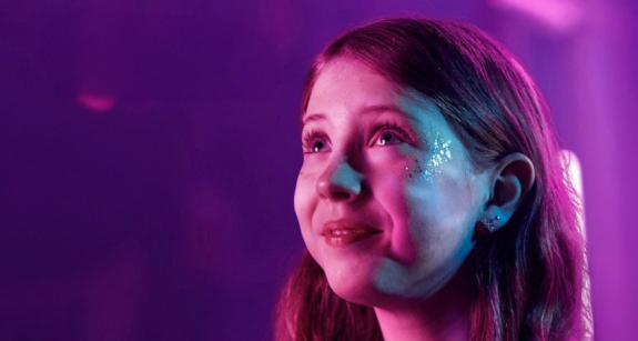 A girl with glitter on her face, possibly at a concert, looks up with a smile. The image has a purple and pink color scheme