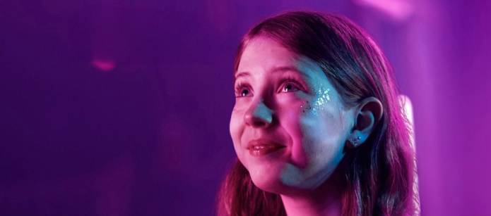 A girl with glitter on her face, possibly at a concert, looks up with a smile. The image has a purple and pink color scheme