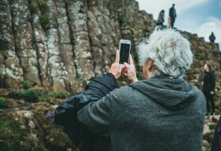 An elderly person with white hair, seen from behind, is holding a smartphone up to take a picture of a rocky, cliff-like formation. Other people can be seen in the distance, climbing or standing on the rocks.