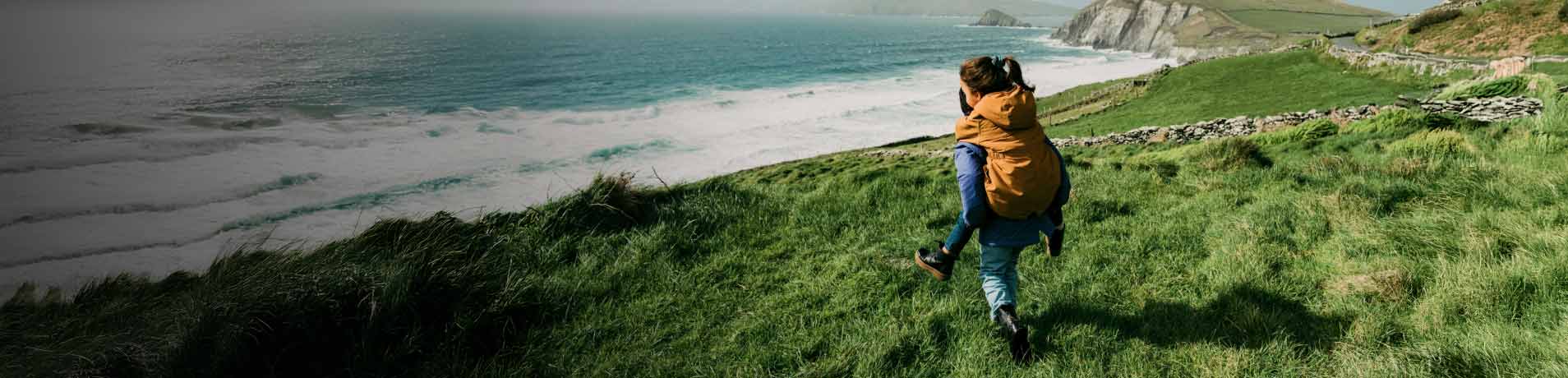 A person gives another a piggyback ride on a green grassy hill overlooking the ocean. Waves are crashing on the shore below, and a misty, green coastline stretches into the distance.