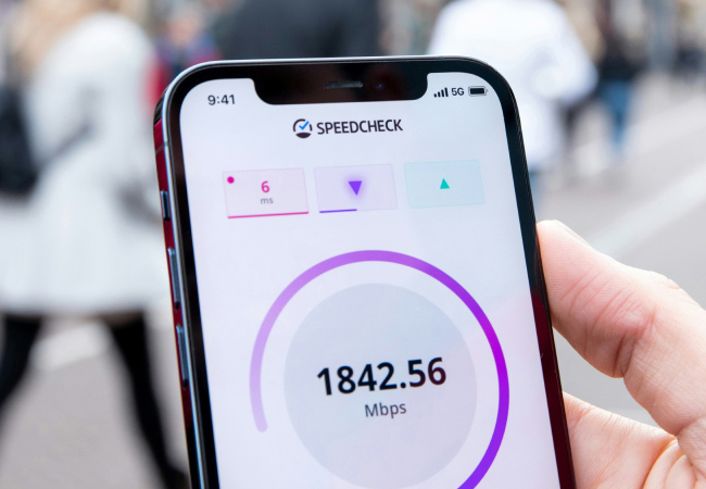 Close-up shot shows a hand holding a smartphone. A speed test app shows a download speed of 1842.56 Mbps and a latency of 6 ms. The phone's status bar indicates a 5G connection. A blurred, urban background is visible behind the hand.