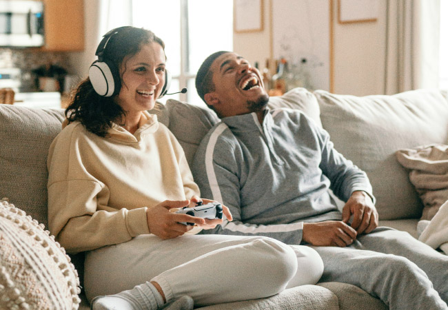 A couple is sitting on a couch, laughing and playing a video game. A woman on the left is wearing a headset with a microphone and is holding a white controller. A man on the right is laughing while sitting next to her.