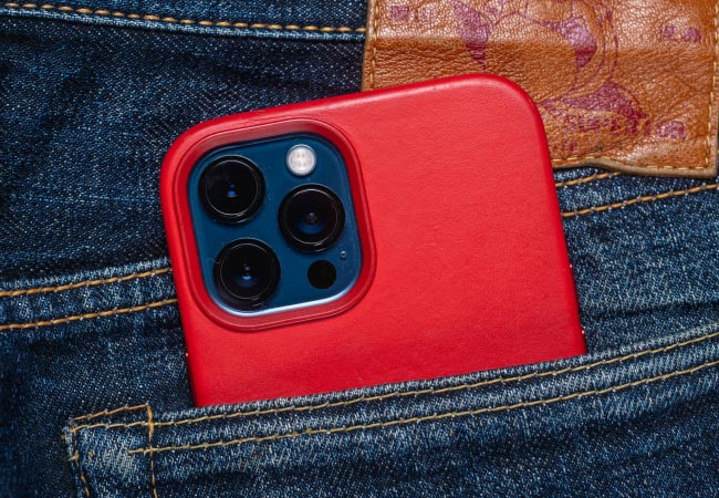 A smartphone, in a vibrant red case, is in the back pocket of a pair of blue denim jeans. The camera lenses are clearly visible, and a leather patch is seen above the pocket.