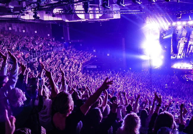 A crowd fills a large, dark concert arena with their hands raised in the air. Bright stage lights and a large video screen are visible on the right side, illuminating the space with a vivid purple and white glow.