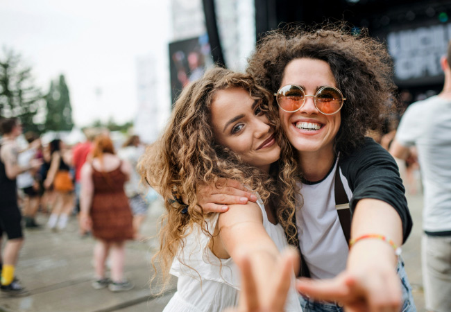 Two smiling friends are posing for a close-up photo at an outdoor event or music festival. One person has their arm wrapped around the other. Both are looking at the camera, and one is making a peace sign with their fingers. A crowd and a stage are visible in the blurry background.