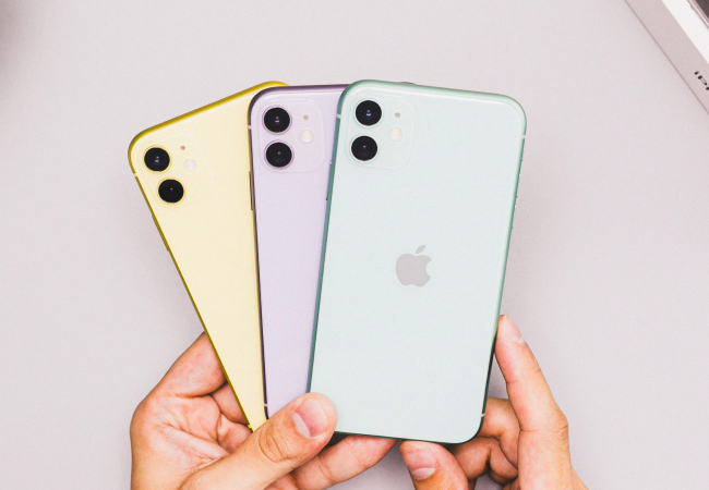 A person's hands are holding three iPhones in a fan-like arrangement against a light gray background, they are in different pastel colors: yellow, purple, and mint green.