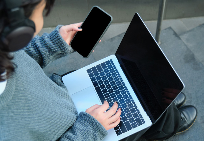 A person, seen from a high angle, is sitting on some stairs and using a laptop. They are also holding a black-screen smartphone in their left hand and wearing headphones. Their right hand is on the laptop's keyboard.