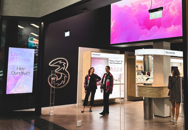 Two people stand in the middle of the Dundrum Three store. The store has a modern design with large digital displays, including a prominent pink cloud-themed screen and another screen advertising "Live @ Three" with the text "Hey Dundrum."