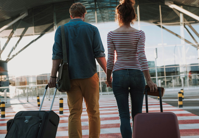 A couple, seen from behind, is walking hand-in-hand toward an airport terminal on a sunny day. They are pulling two suitcases behind them on a striped crosswalk.
