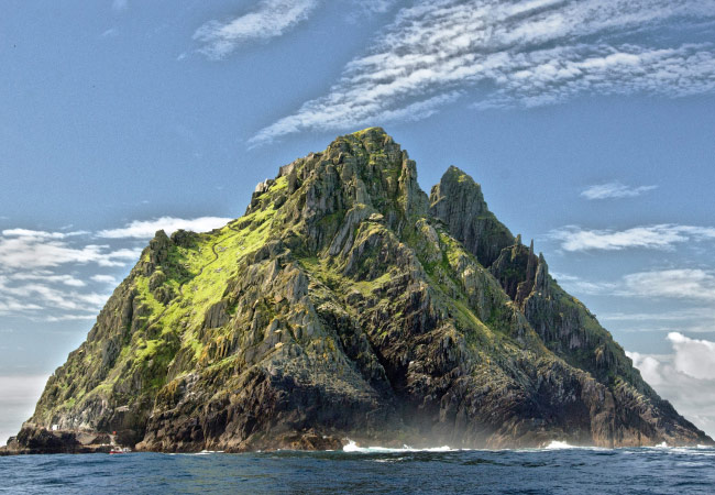 A rugged, rocky island with steep, jagged cliffs juts out of the ocean. The upper half is covered in vibrant green grass and patches of rock. Waves hitting the rocks and the sky is a bright blue with wispy white clouds.