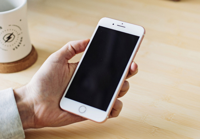 A person is holding a white smartphone with a black, blank screen in their left hand. A white mug is out of focus in the background, resting on a light-colored wooden table.
