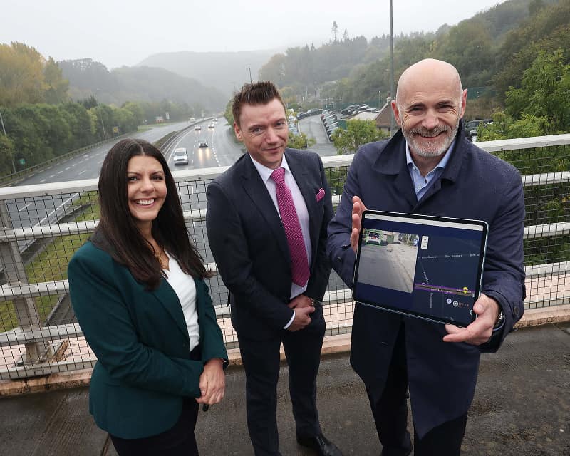 Eoin McManus poses with two people spearheading the new Emerald Motorway Corridor while standing on a motorway bridge overlooking the road, while holding up an iPad showing the proposed digital corridor