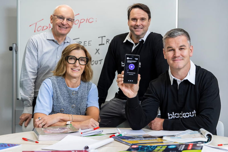 Johnny Sexton holding up a smartphone alongside three colleagues gathered around a desk with educational materials, in front of a whiteboard.