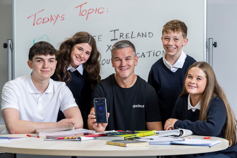 Johnny Sexton holding up a smartphone alongside four smiling school students gathered around a table with schoolbooks, in front of a whiteboard reading "Today's Topic: Three Ireland x Blackout Education.