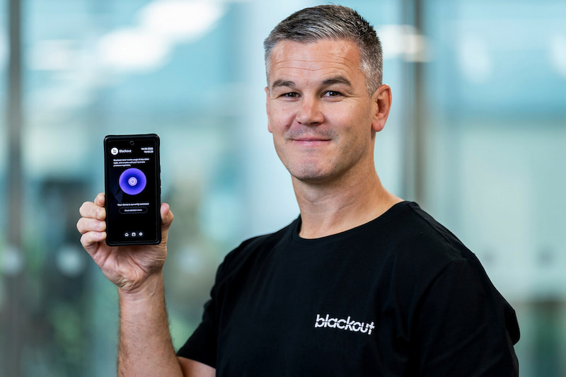 Former Irish rugby captain Johnny Sexton holding up a smartphone displaying the Blackout app interface.