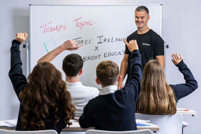 Johnny Sexton standing in front of a whiteboard reading "Today's Topic: Three Ireland x Blackout Education" as students raise their hands in a classroom.