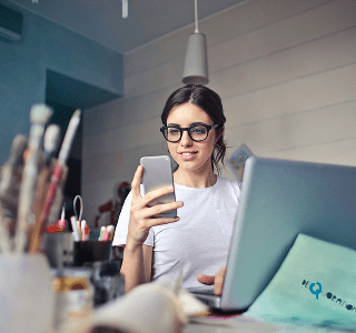 A woman wearing a white t-shirt and black glasses sitting in front of her laptop on a messy desk and looking at her phone