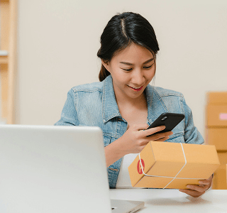 A woman wearing a jean jacket sitting at a desk in front of her laptop while taking a picture of a brown box with her phone