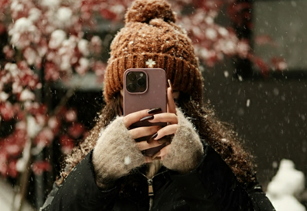 Person with curly hair and a brown knit hat taking a photo with a smartphone in the snow.